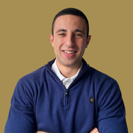 Un uomo con i capelli corti e scuri sorride, indossa un pullover blu e una camicia bianca con colletto, in posa su un semplice sfondo marrone.
